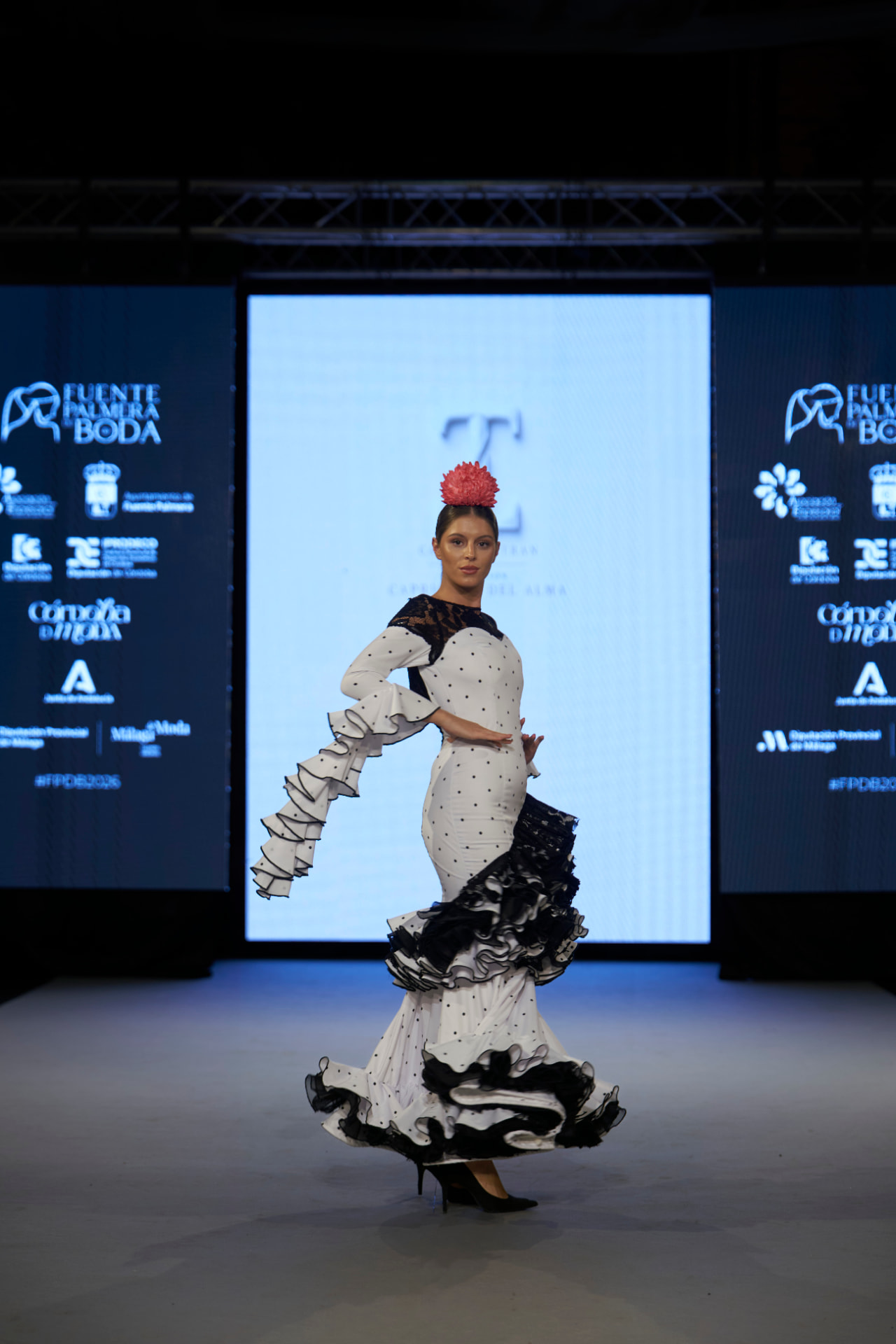 Traje de flamenca blanco de lunares con volante asimétrico negro, posando con el brazo extendido